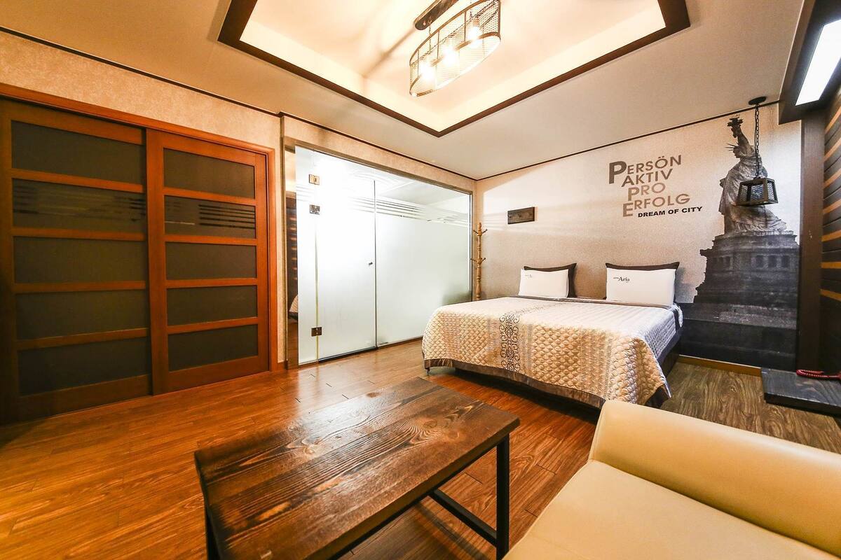 A hotel room with a bed, a wooden table, a sofa, a wardrobe, and a wall with a statue and text.