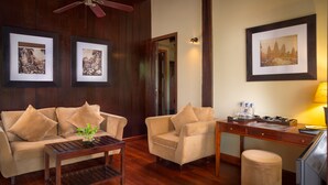 A room with a wooden ceiling fan, a sofa with cushions, a coffee table, a desk with a lamp, and framed pictures on the wall. - GZ Eden Privilege Resort and Spa (Siem Reap)