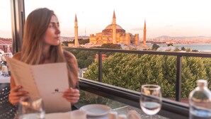 A woman reading a menu at a table with a view of a historic building and a river. - Sura Design Hotel and Suites - Boutique Class (Istanbul)