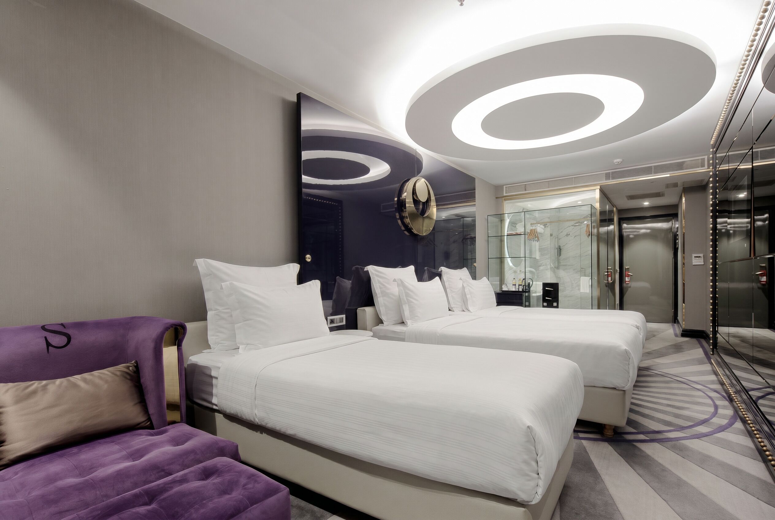 Una habitación de hotel moderna con dos camas, un sofá púrpura y un baño con paredes de vidrio.