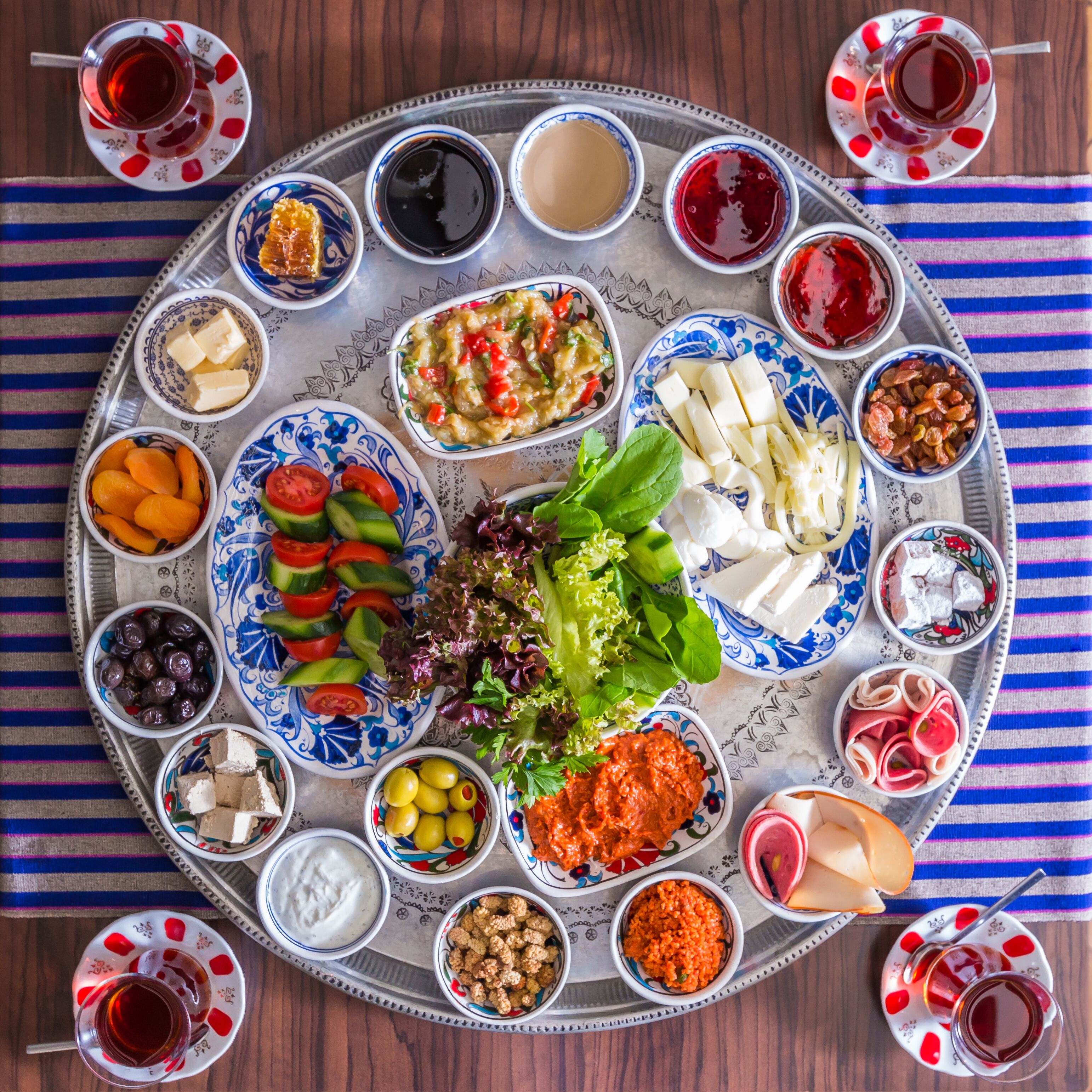 A large, round, silver tray with various food items and drinks, including cheeses, olives, and a salad, arranged on a striped cloth.