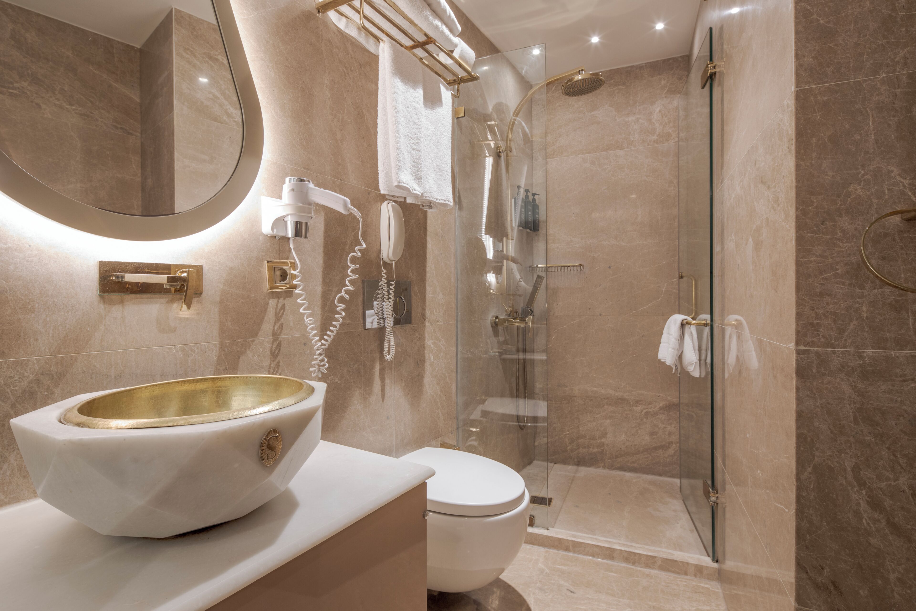 A bathroom with a large mirror, a white sink with a gold basin, a toilet, and a glass-enclosed shower area.