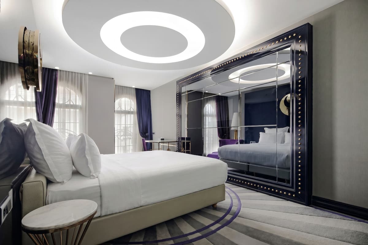 Une chambre d’hôtel moderne avec un grand lit, un grand miroir rond à cadre noir, une petite table ronde et un plafond orné de luminaires circulaires.