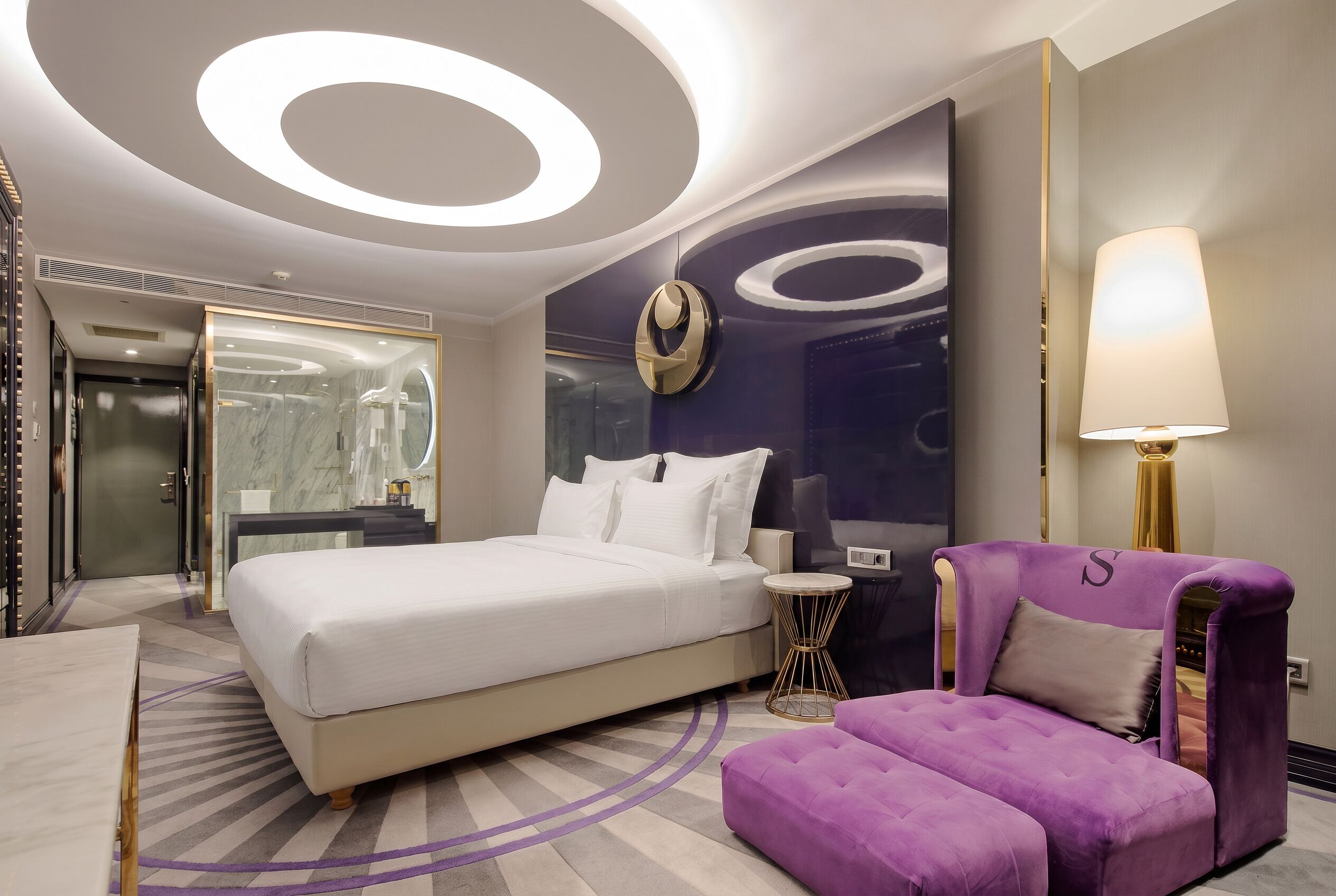 Une chambre d’hôtel moderne avec un grand lit, un fauteuil violet, une table ronde et une lampe fixée au mur.