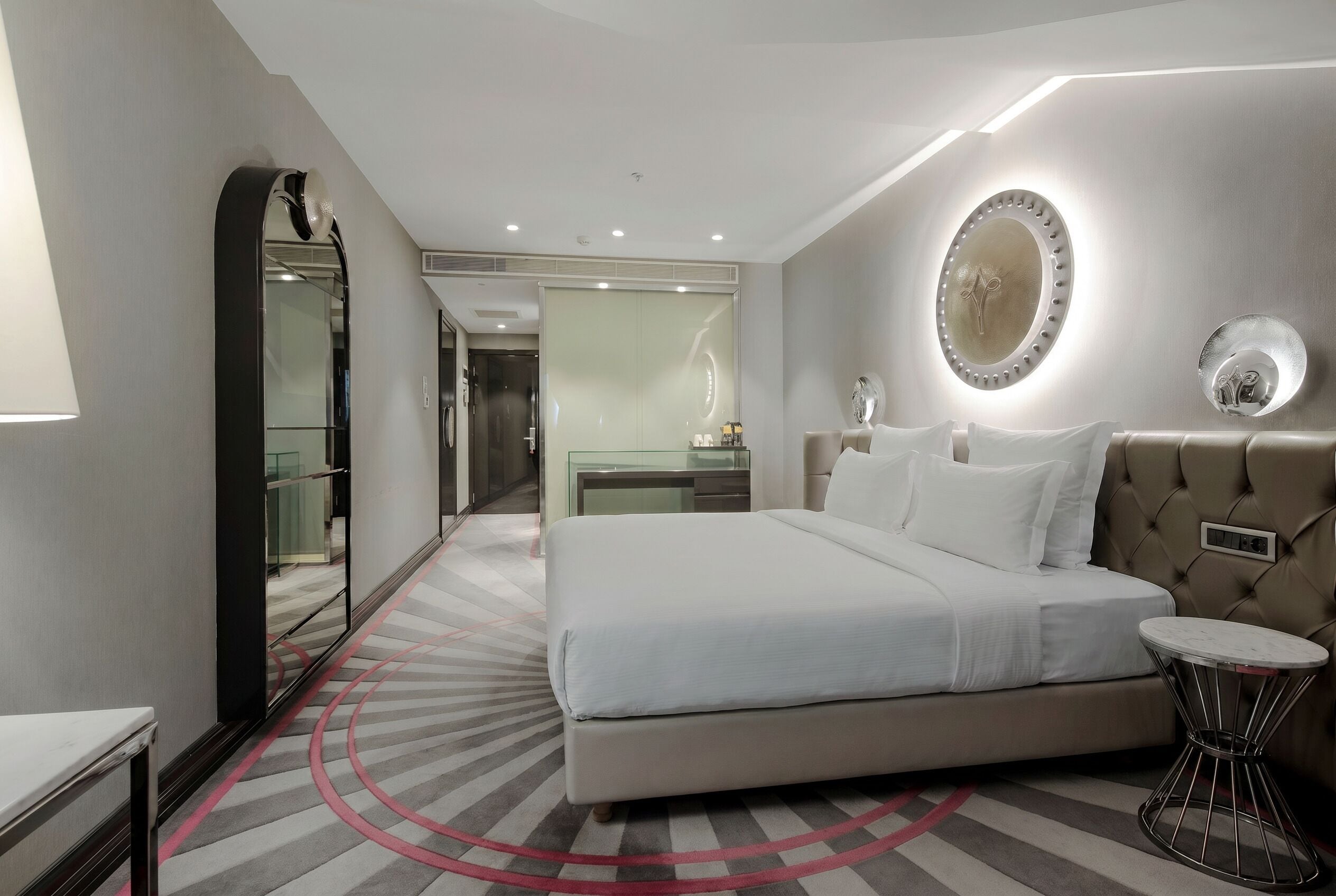A modern hotel room with a large bed, a round mirror, a bedside table, and a bathroom visible in the background.