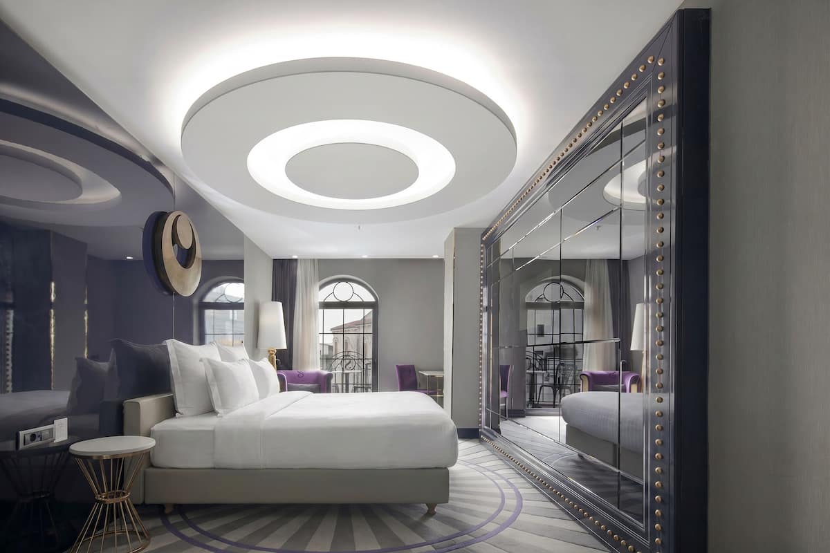 Une chambre d’hôtel moderne avec un grand lit, un luminaire rond au plafond, un miroir orné d’un motif de perles et une vue sur la ville grâce à de grandes fenêtres.