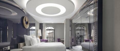 A modern hotel room with a large bed, a round ceiling light, a mirror with bead trim, and a view of the city through large windows.