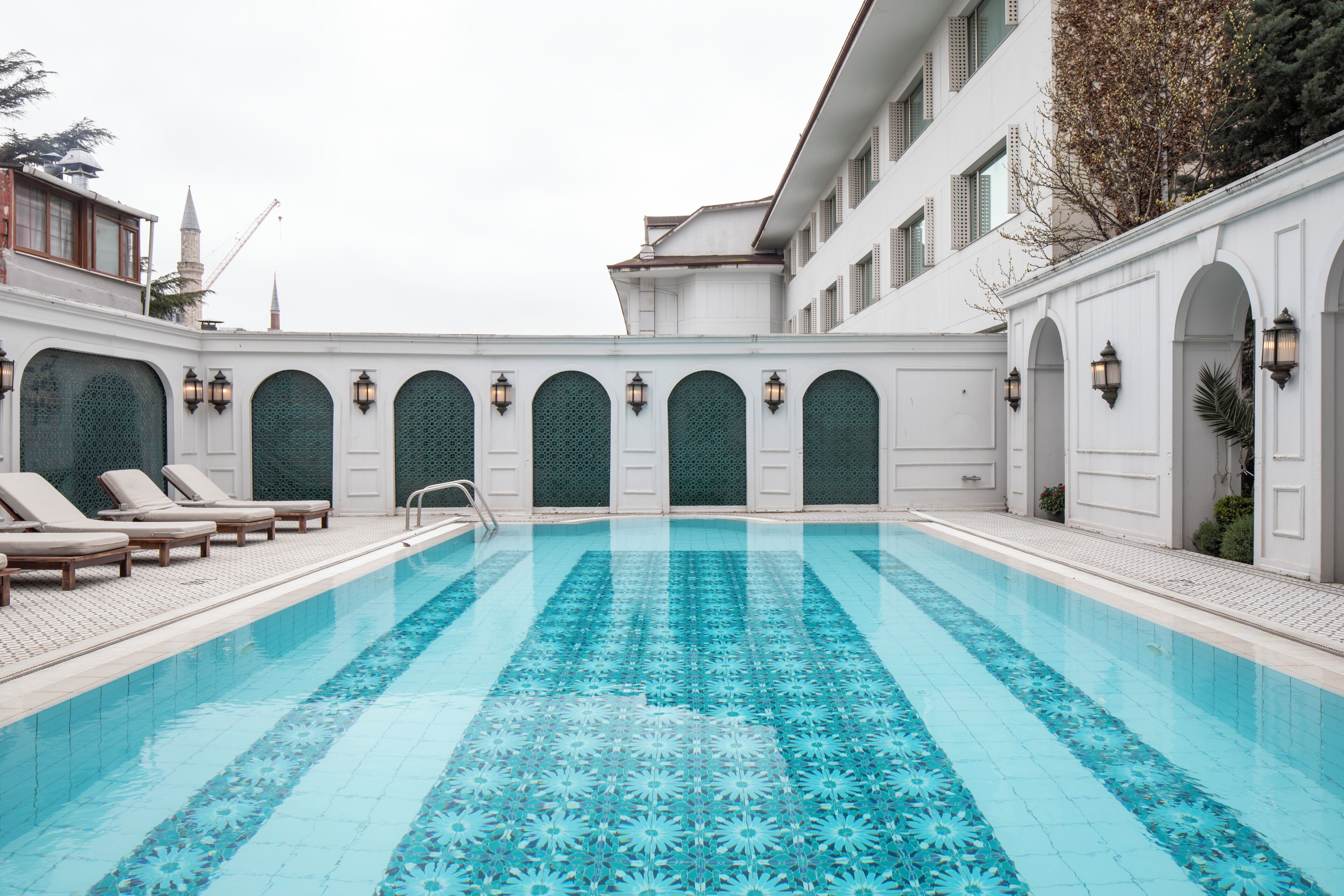 Ein Schwimmbecken mit geometrischem Fliesenmuster, umgeben von weißen Wänden mit Rundbogenöffnungen und Wandleuchten.