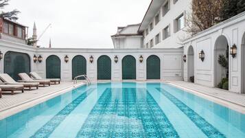 Ein Schwimmbecken mit geometrischem Fliesenmuster, umgeben von weißen Wänden mit Rundbogenöffnungen und Wandleuchten.