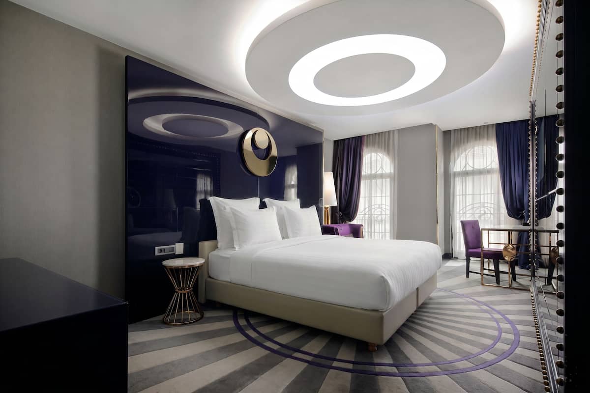 Une chambre d’hôtel moderne avec un grand lit, un luminaire rond au plafond, une petite table ronde et une chaise.