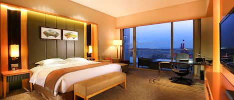 Habitación de hotel con una cama grande, un escritorio, una silla y vistas de la ciudad a través de amplias ventanas.