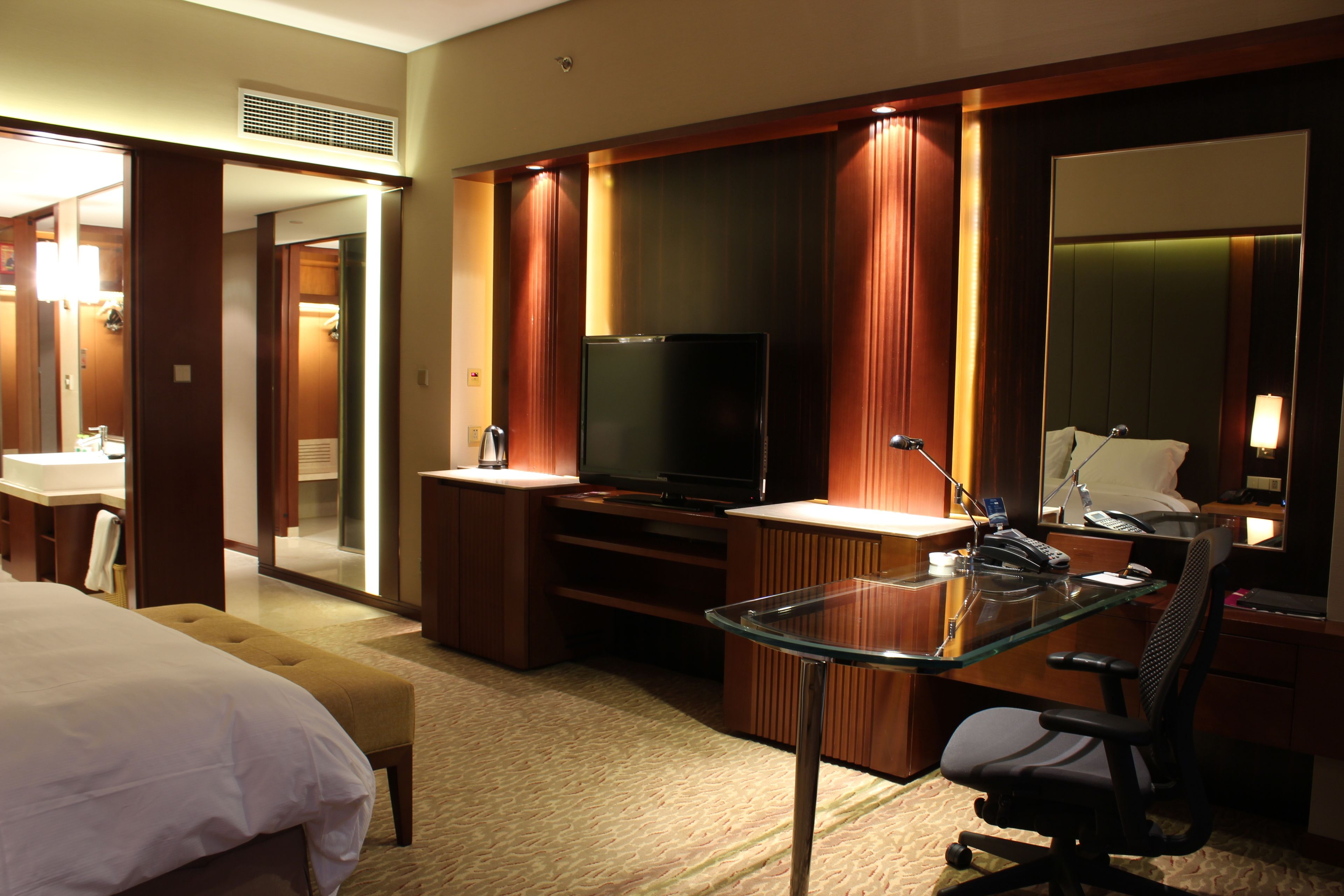 A hotel room with a bed, a glass desk, a television, and a bathroom.