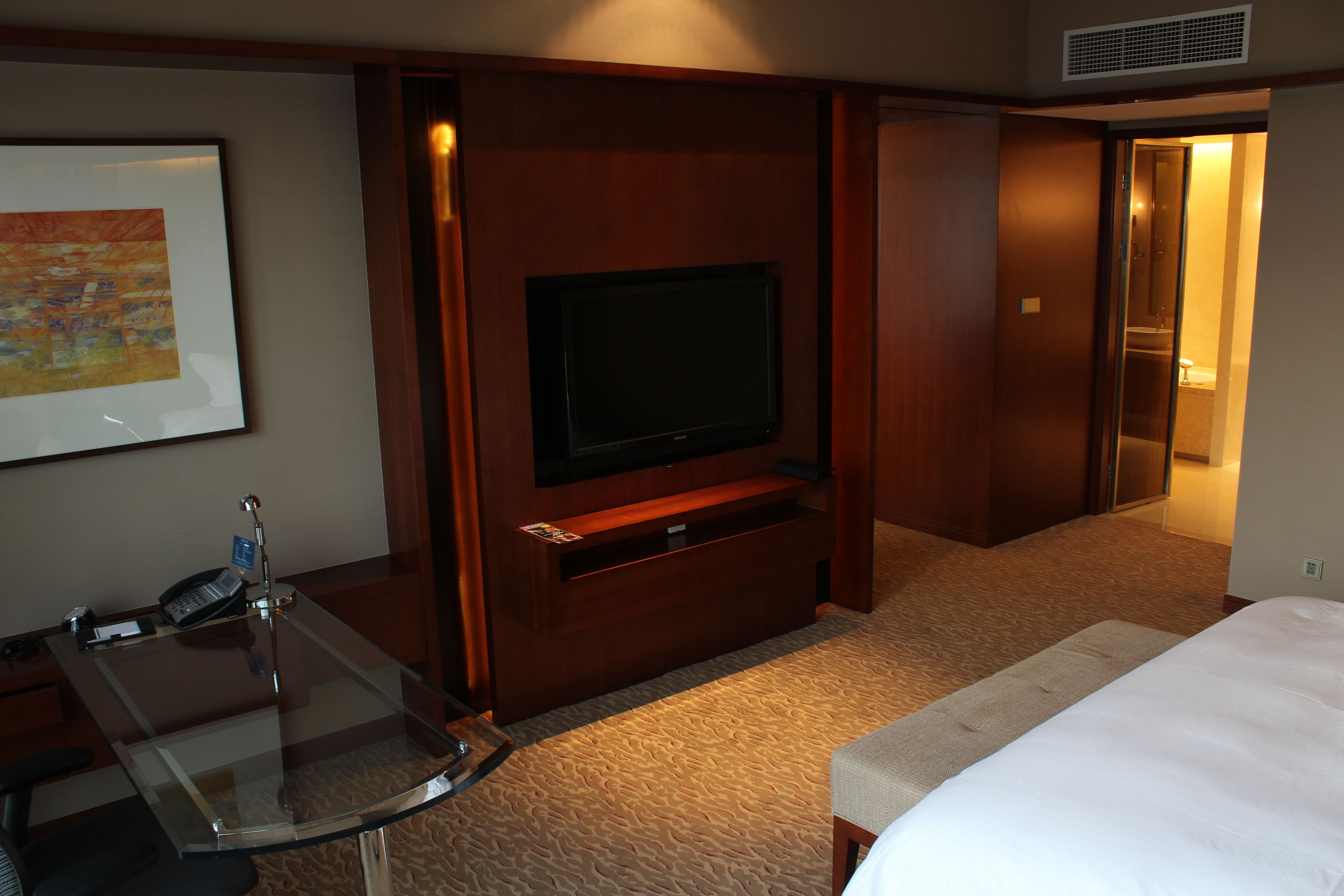 A hotel room with a bed, a TV mounted on a wooden panel, a glass desk with a phone, and a carpeted floor.