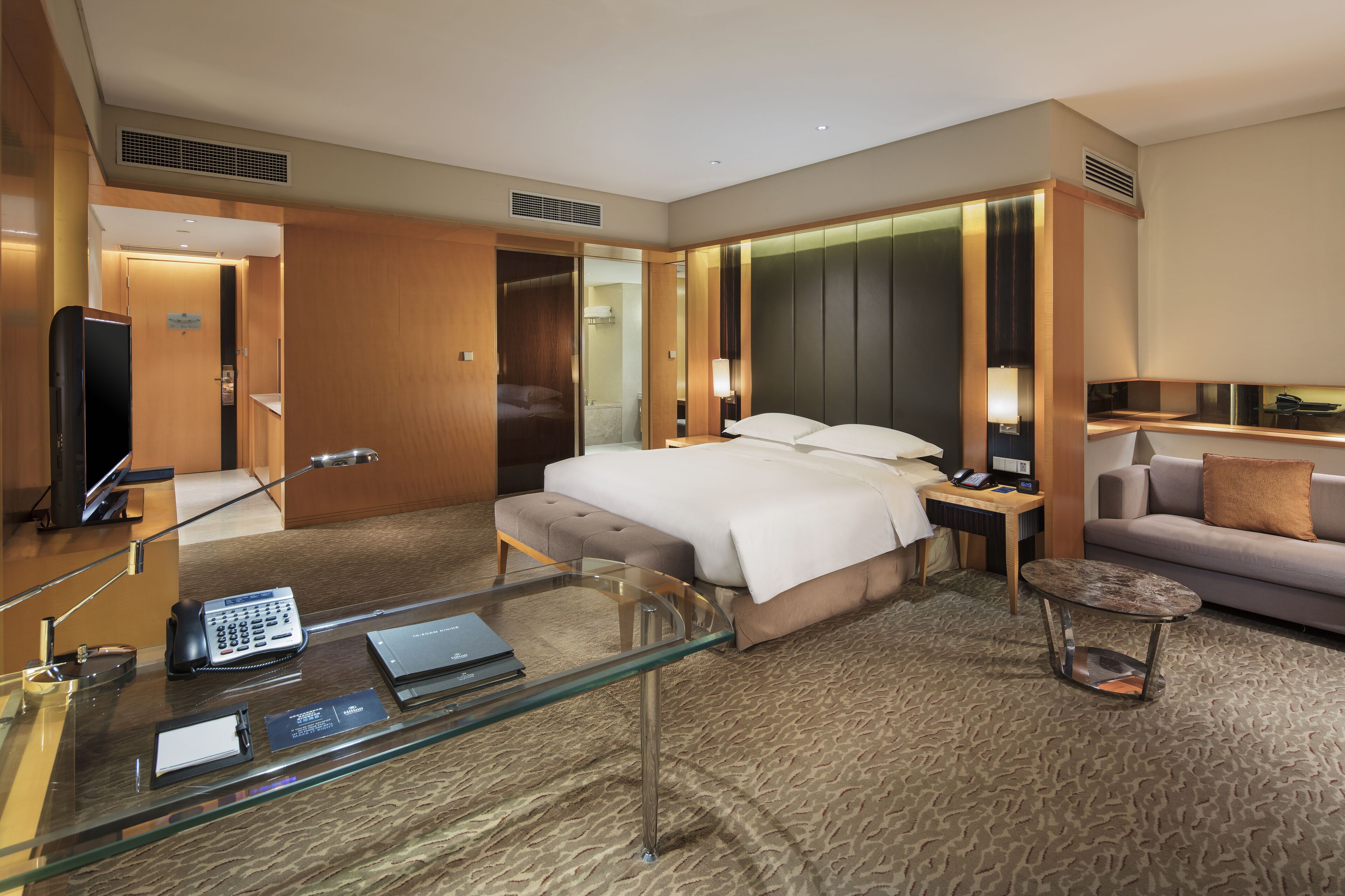 A hotel room with a large bed, a glass coffee table, a sofa, a desk with a telephone, and a television.