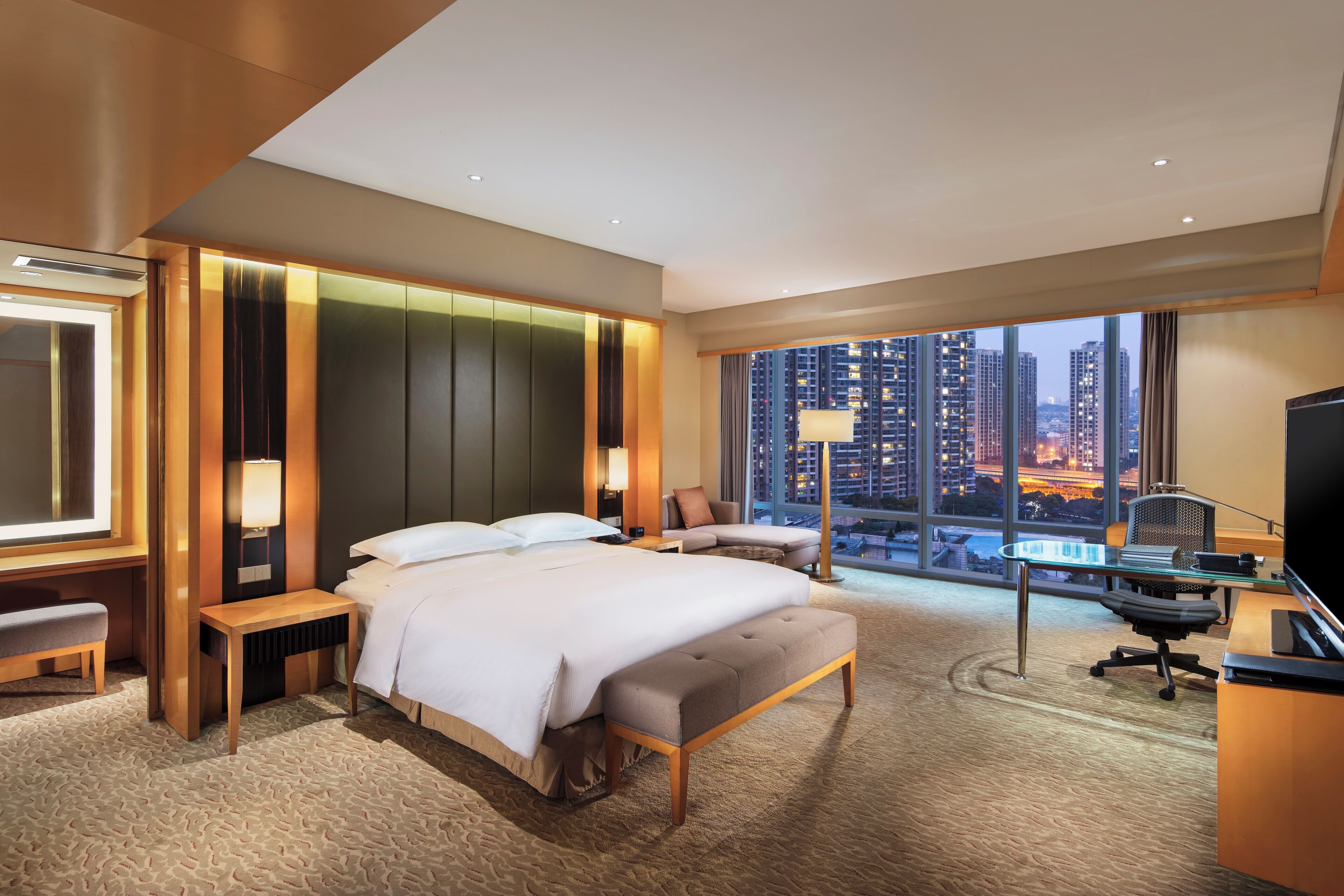 A hotel room with a large bed, a desk with a chair, a TV, and a view of a cityscape.