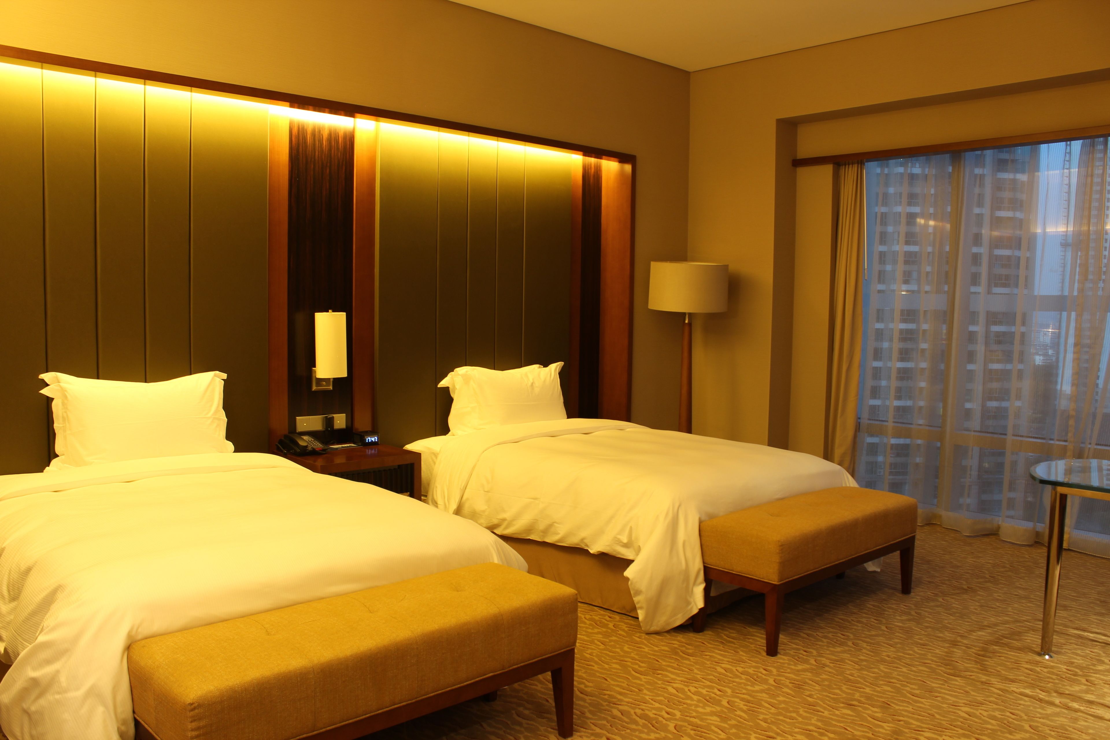 A hotel room with two beds, a bedside table with a lamp, a small table, and a large window with curtains.
