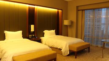 A hotel room with two beds, a bedside table with a lamp, a small table, and a large window with curtains.