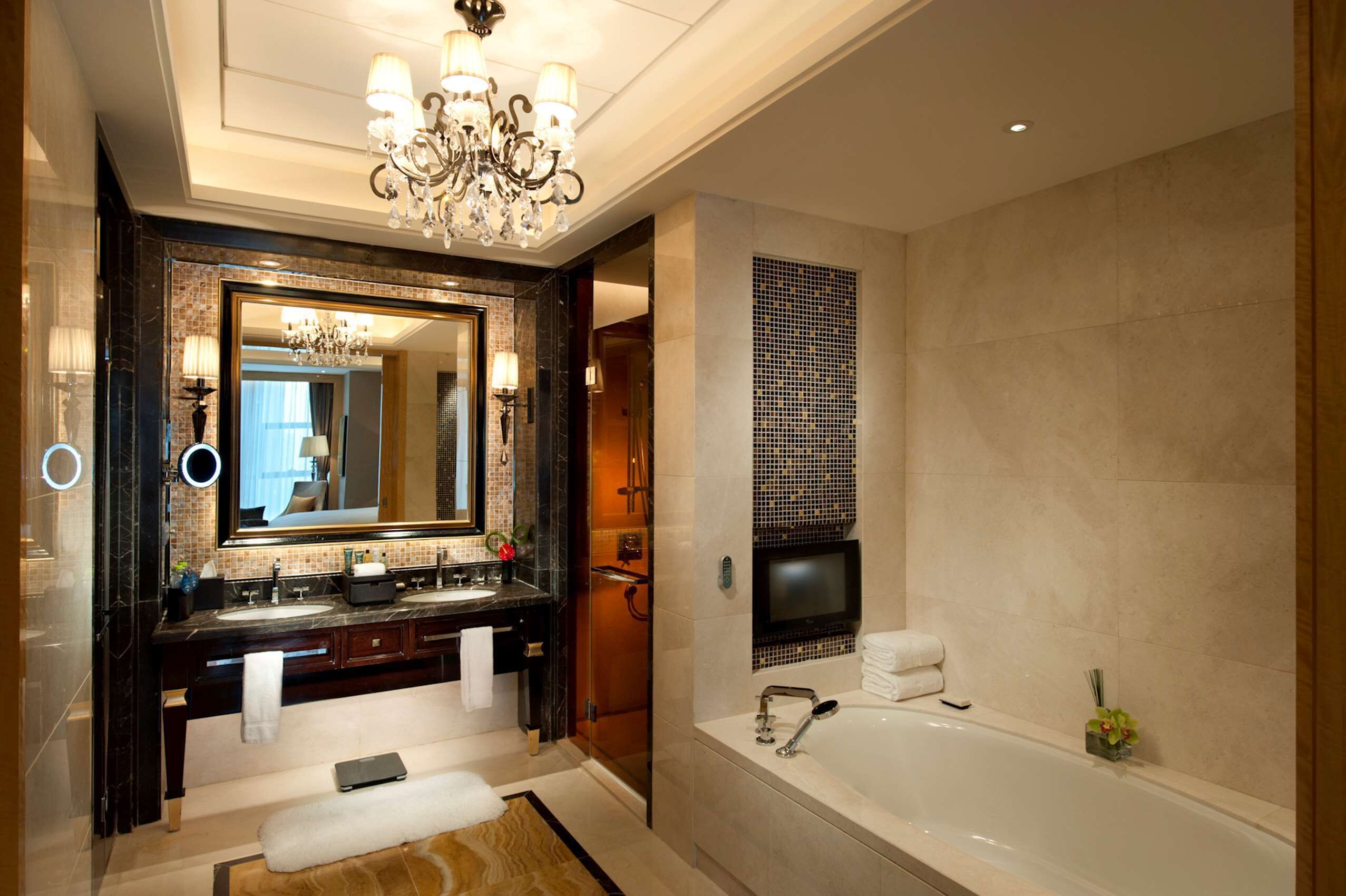 A luxurious bathroom with a large mirror, a chandelier, a bathtub, and a vanity with a television.