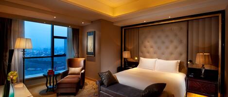 A hotel room with a large bed, a sofa, a chair, a desk, and a view of the city through large windows.