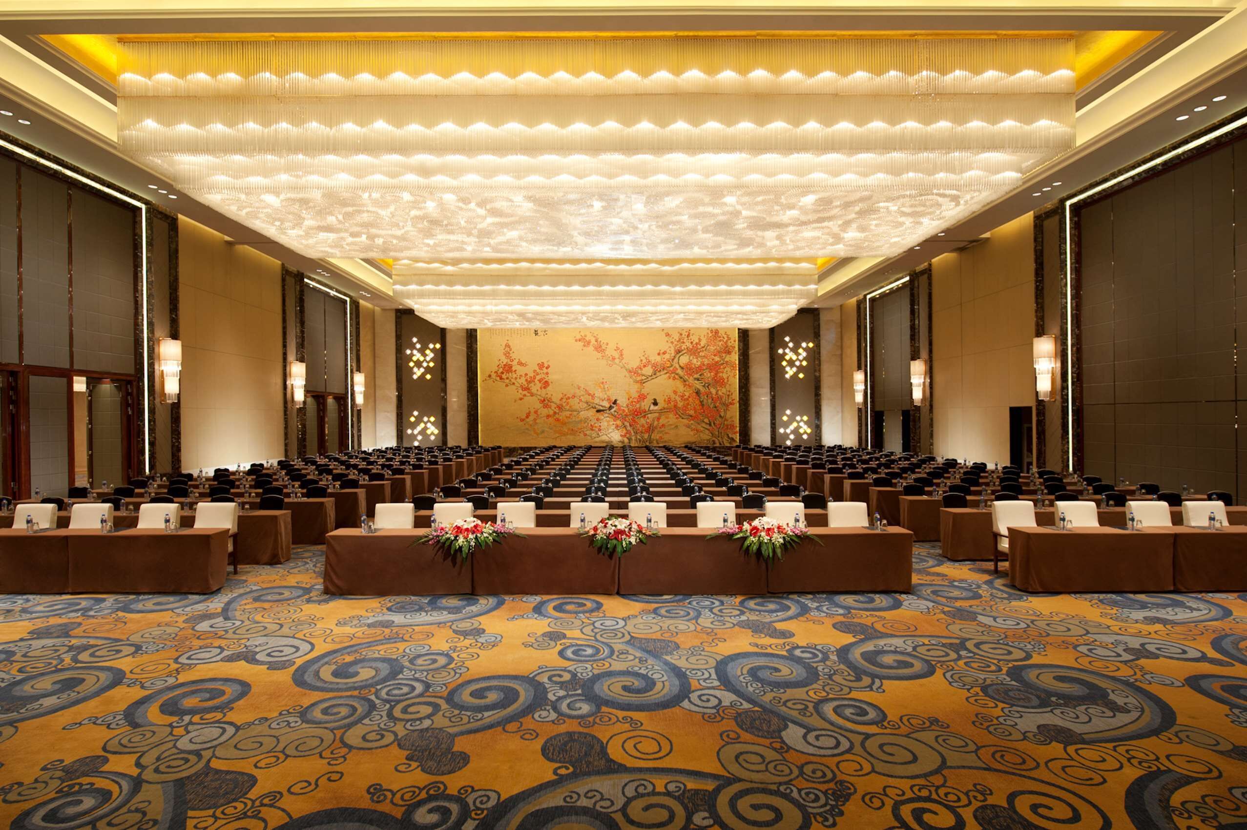 Une salle de conférence avec des rangées de tables, une moquette à motifs et une grande fresque florale sur le mur du fond.