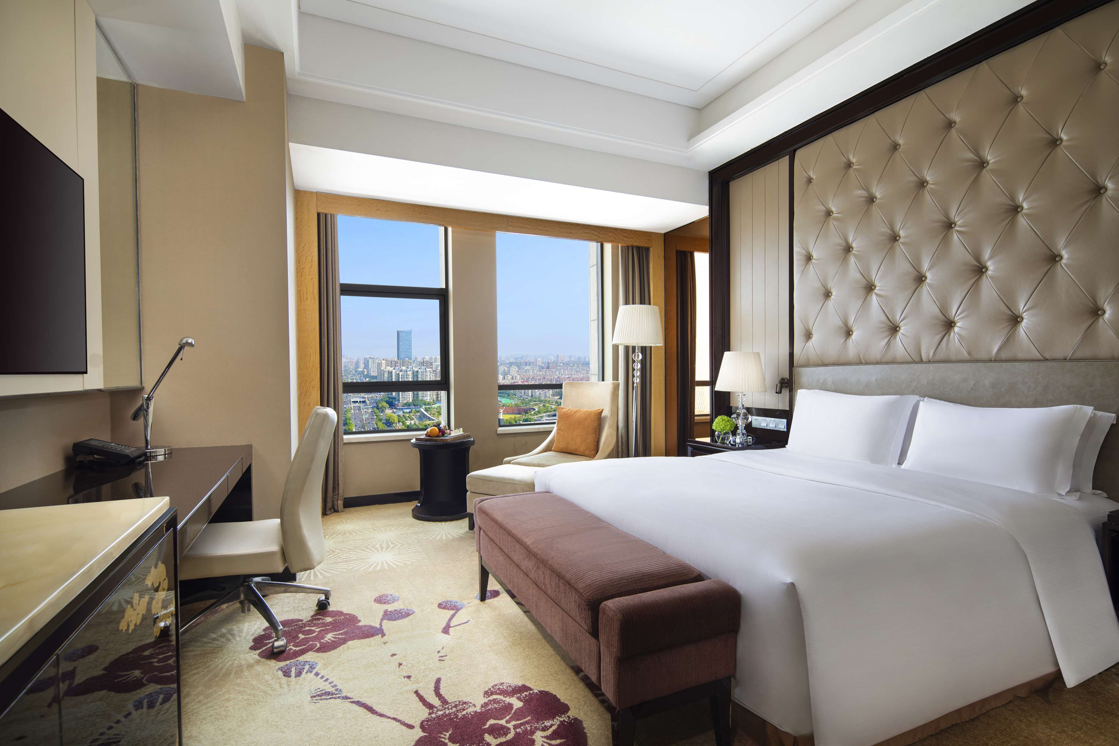 A hotel room with a large bed, a desk, a chair, a TV, and a view of the city.