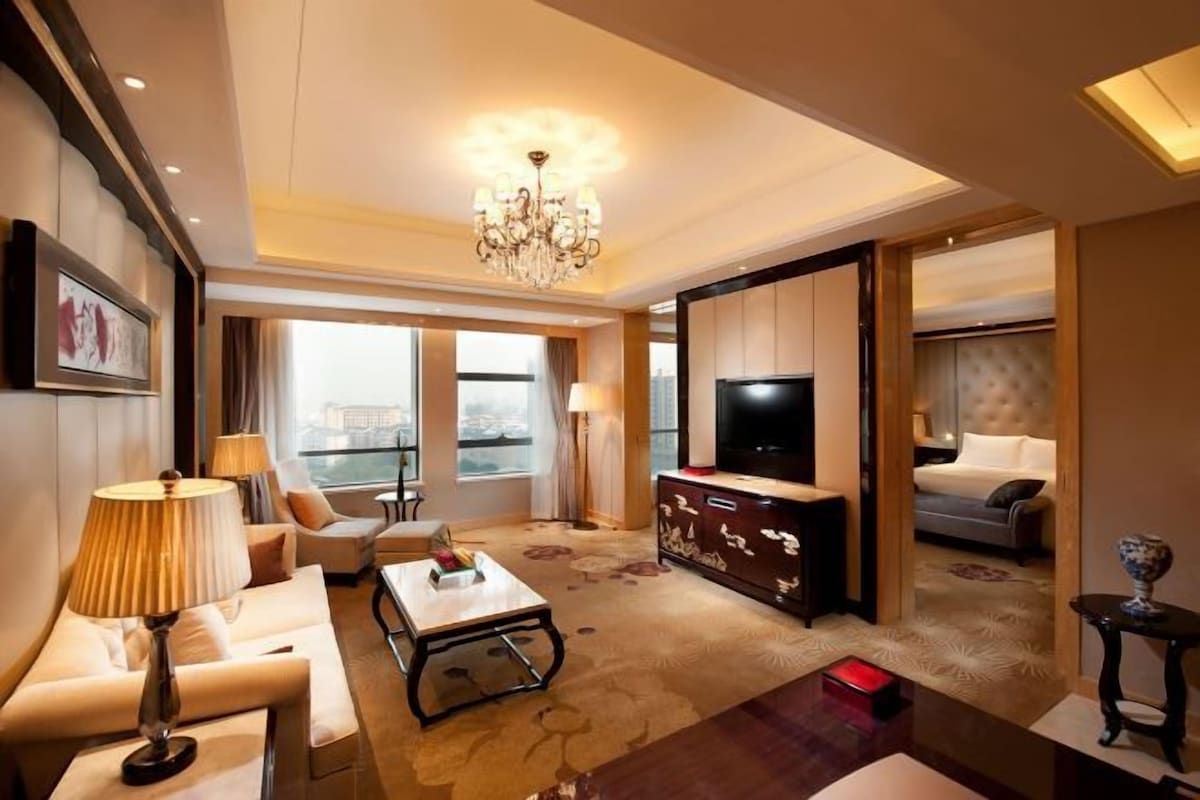 A spacious hotel room with a large bed, a TV, a seating area with a coffee table, and a view of the city.