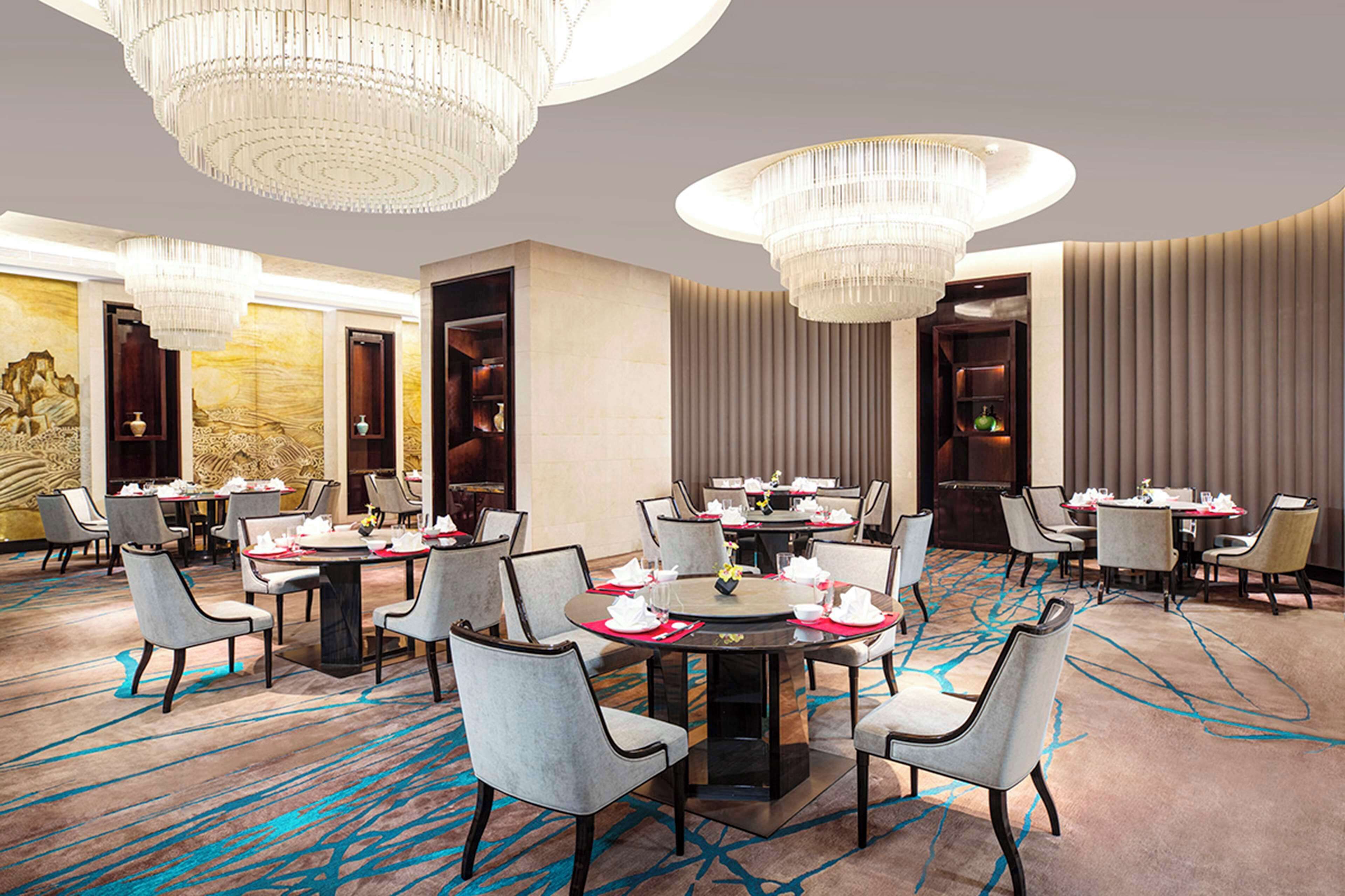 Ein Hotelrestaurant mit mehreren runden Tischen, grau gepolsterten Stühlen und großen Kristalllüstern.