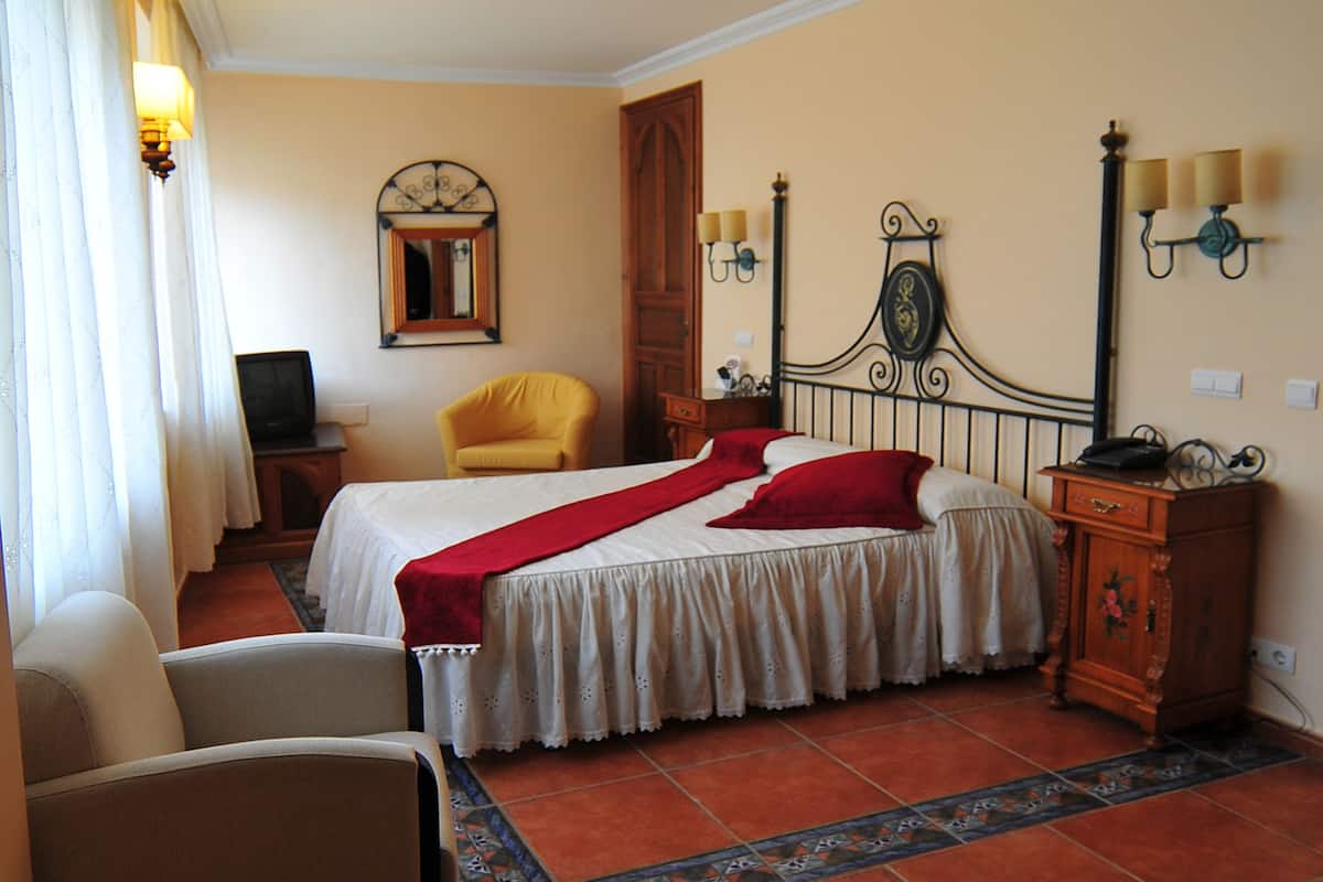 A hotel room with a bed, two chairs, a nightstand, a mirror, and wall sconces.