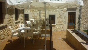 Een buitenruimte met witte tafels en stoelen, een paraplu met AMSTEL branding, en stenen muren.