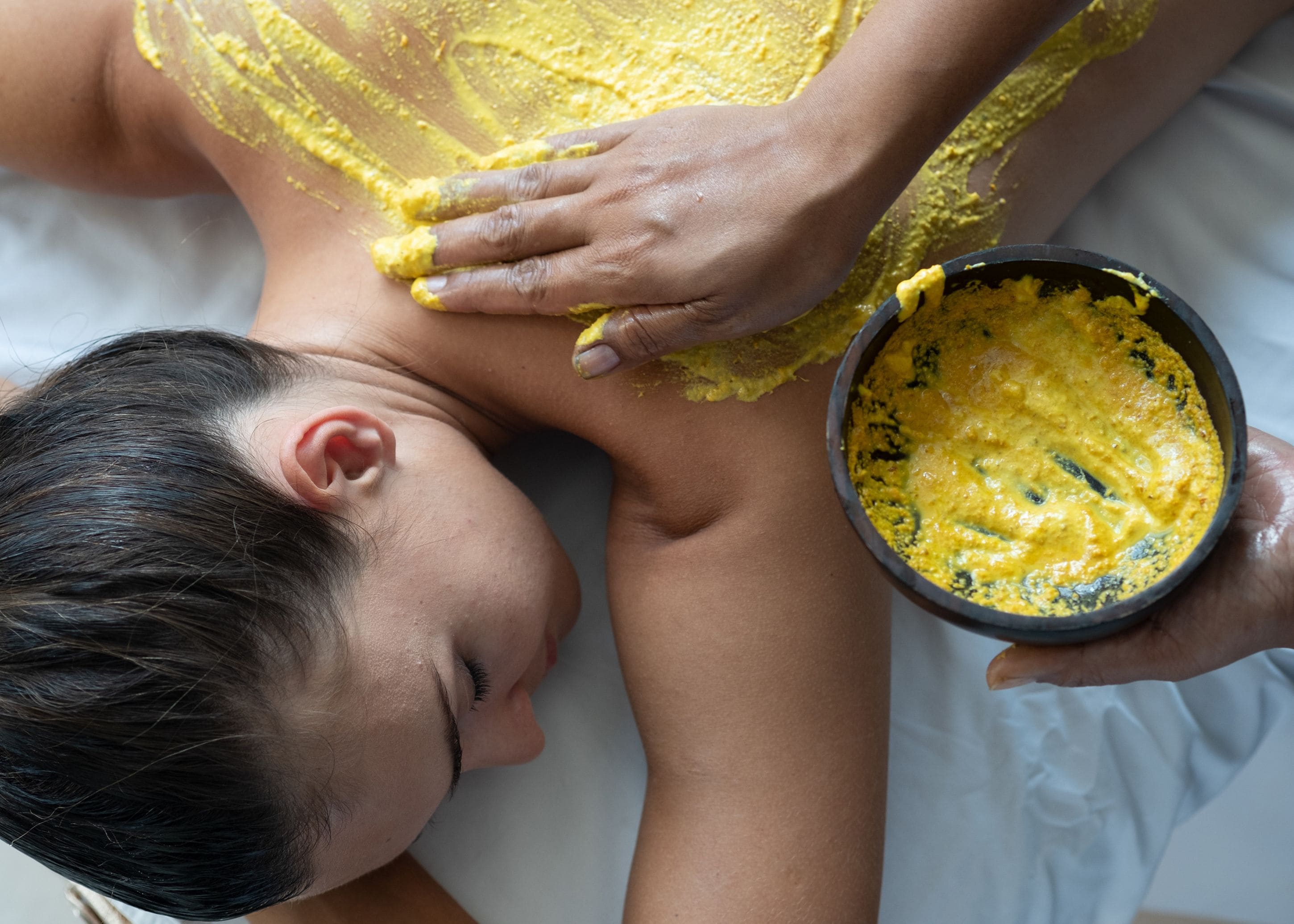A person lying on a bed with a bowl of yellow substance on their arm.