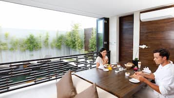 A dining area with a view of a water body and greenery, featuring a wooden table, cushioned seating, and a modern interior design.