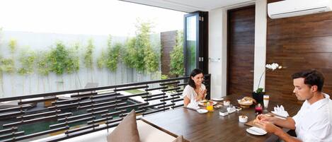 A dining area with a view of a water body and greenery, featuring a wooden table, cushioned seating, and a modern interior design.