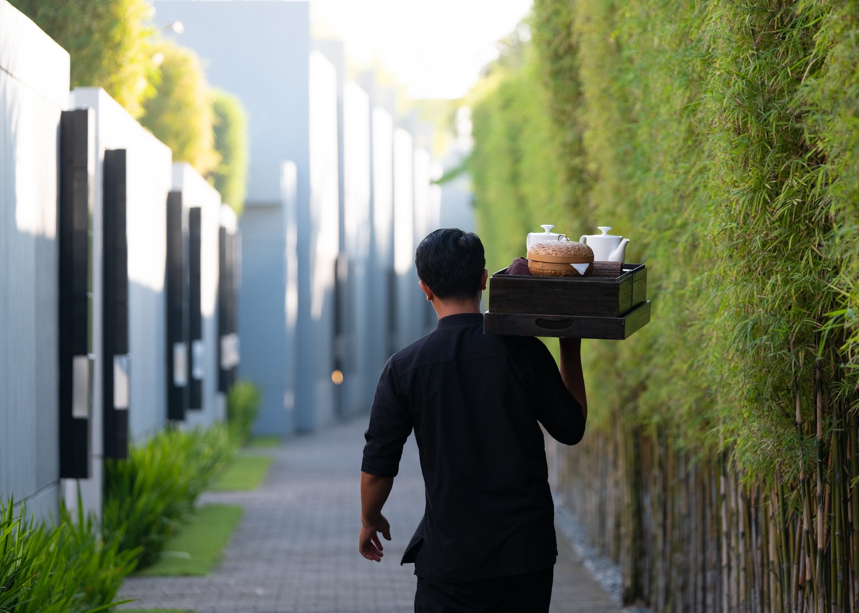 Une personne transporte un plateau rempli d’ustensiles de cuisine le long d’un étroit sentier bordé de verdure et de bâtiments modernes.