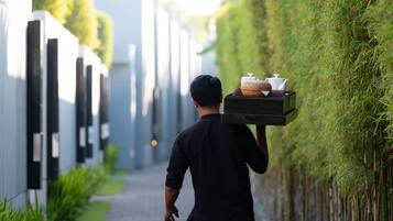 Une personne transporte un plateau rempli d’ustensiles de cuisine le long d’un étroit sentier bordé de verdure et de bâtiments modernes.