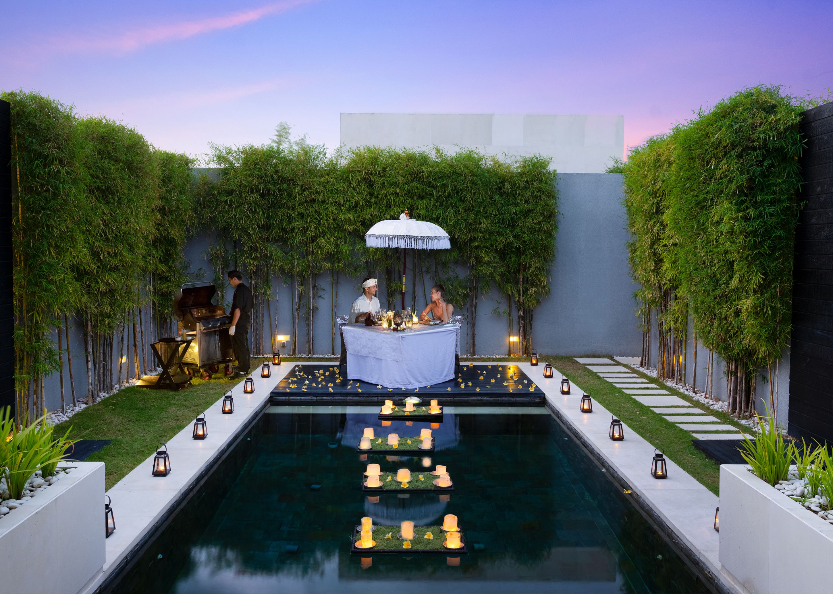 A serene outdoor dining area with a pool, surrounded by bamboo plants and a modern building.