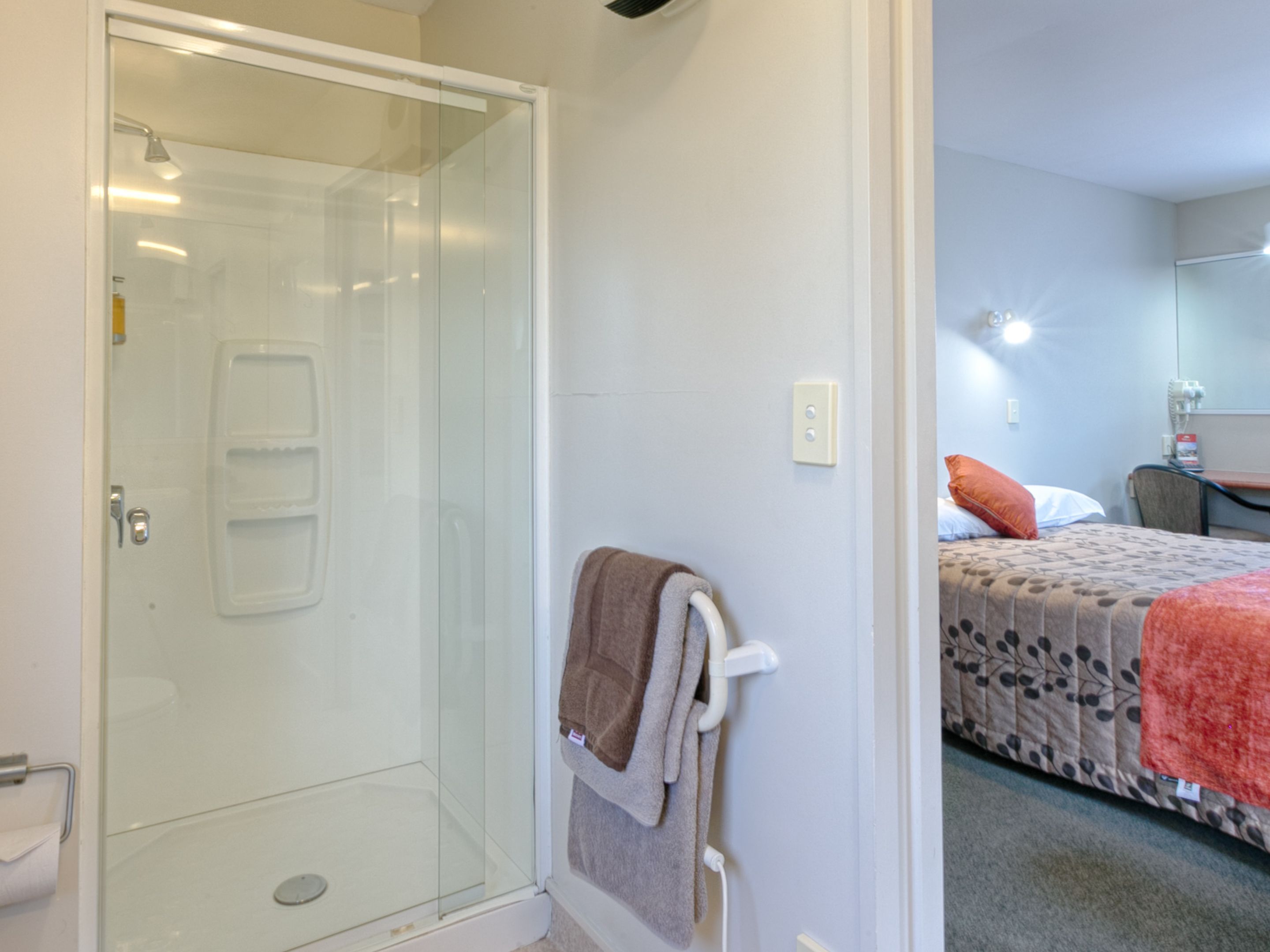 A hotel room with a glass shower enclosure, a towel rack with a towel, a bed with a patterned bedspread, and a small table with a chair.
