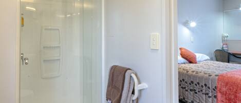 A hotel room with a glass shower enclosure, a towel rack with a towel, a bed with a patterned bedspread, and a small table with a chair.