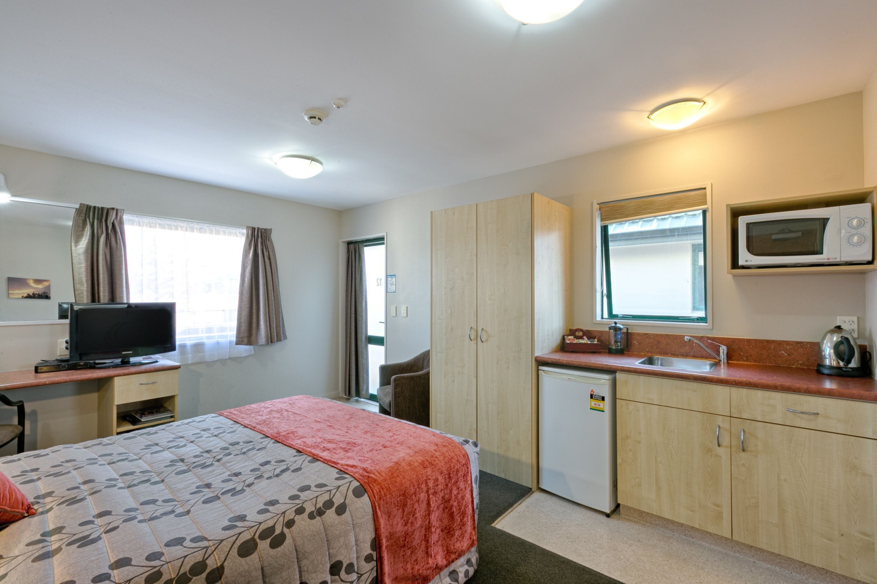 Quarto de hotel com cama, escrivaninha com televisão, cozinha compacta e banheiro.