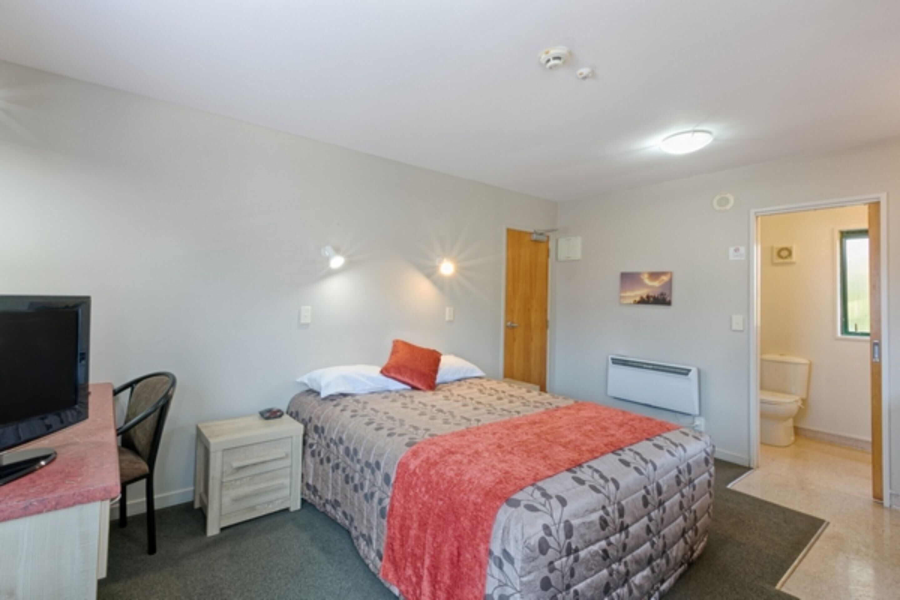Une chambre à coucher avec un lit, un bureau avec un ordinateur, une chaise, une télévision, une porte et une fenêtre.