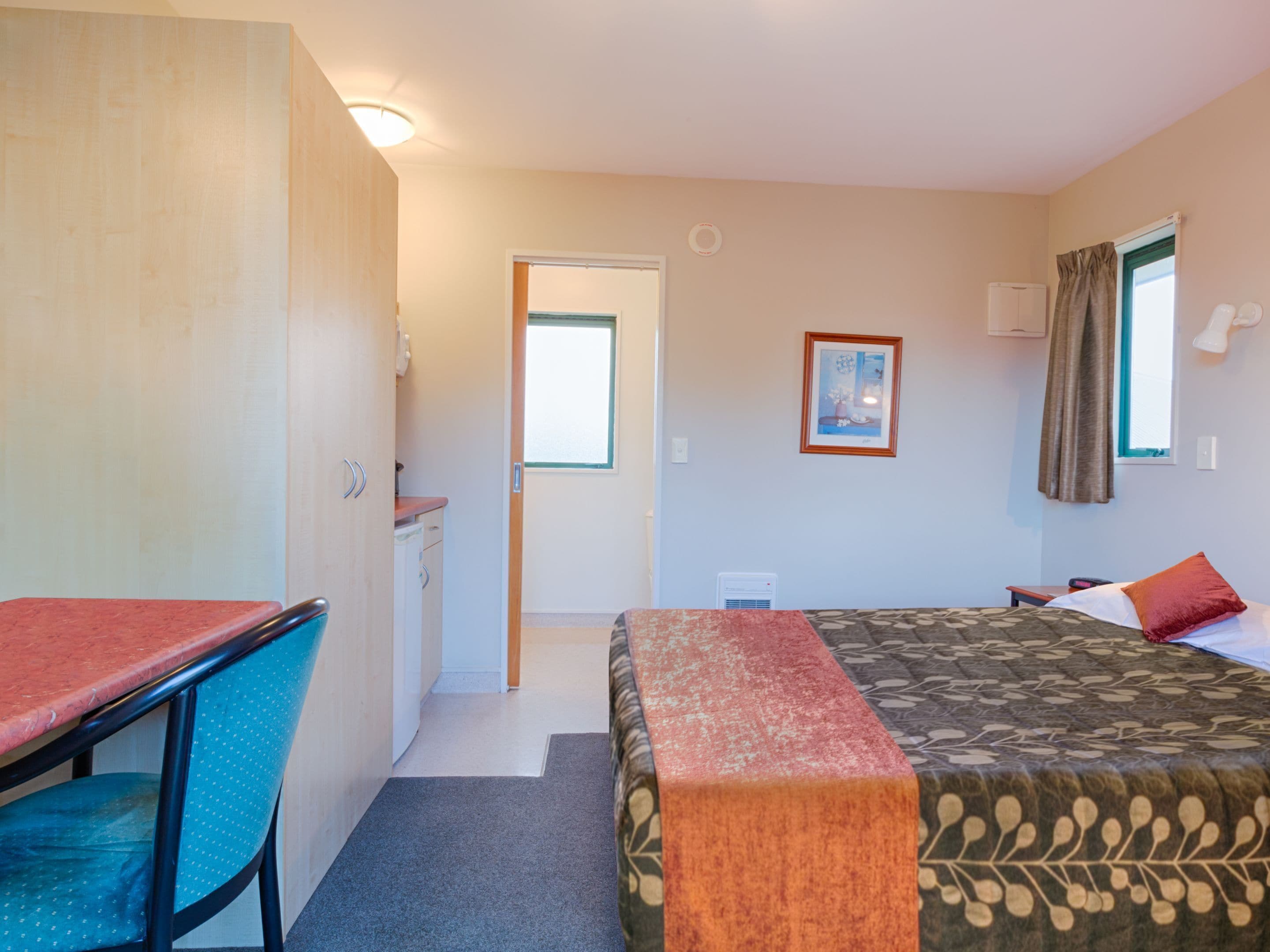 Habitación de hotel con cama, escritorio, silla, kitchenette pequeña y ventana con cortinas.