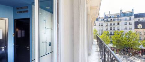 A balcony with a glass door leading to a bathroom, overlooking a street with buildings and people.