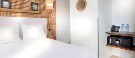 Habitación de hotel con cama, lámpara de pared, cuadro enmarco, adorno de pared tejido, minibar y botella de agua sobre una estantería.