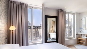 A hotel room with a large bed, a bathroom with a bathtub, and a city view through the windows. - Hôtel Emile Le Marais (Paris)