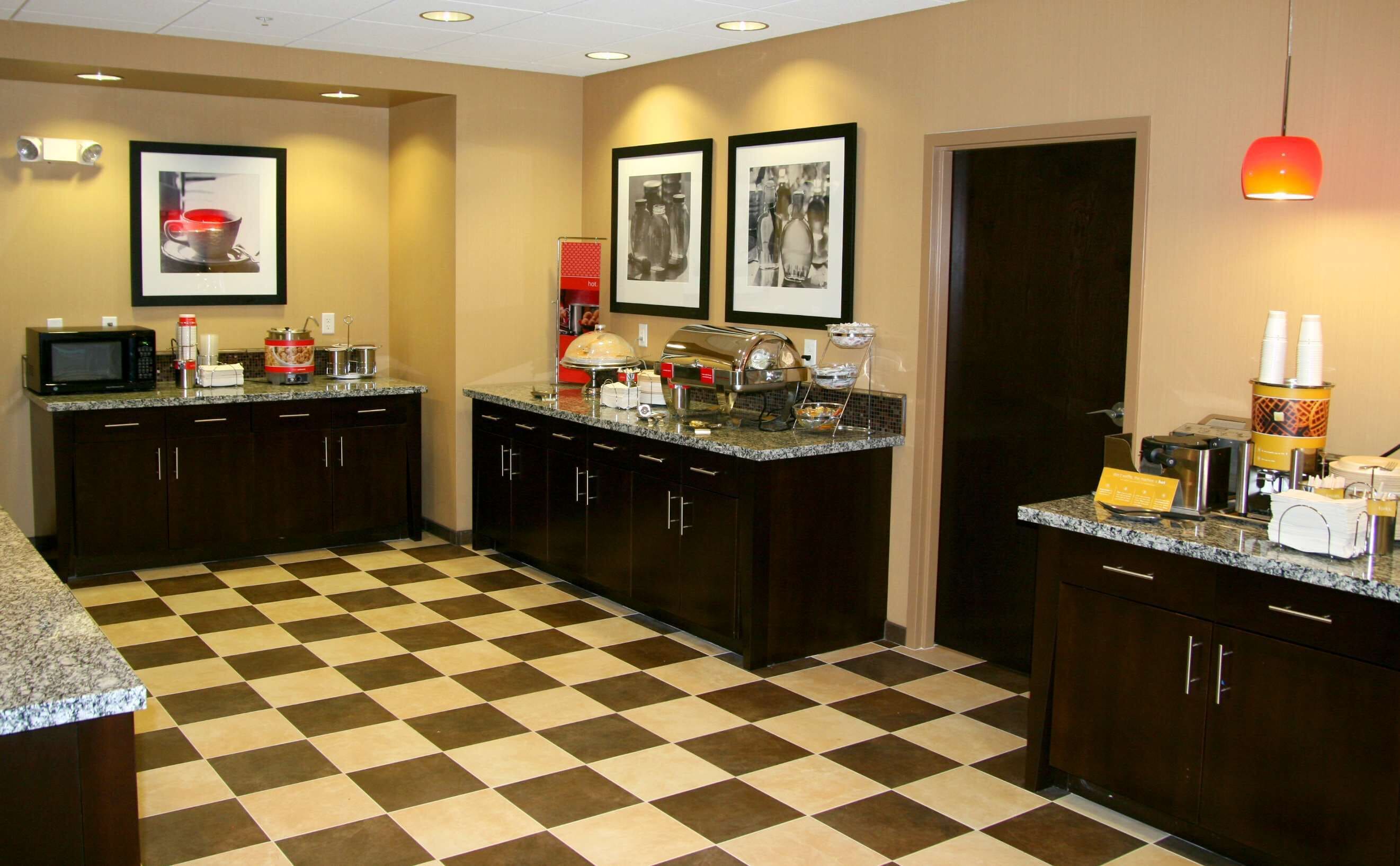 Una cocina de hotel con suelo a cuadros, armarios oscuros, microondas, cafetera y cuadros en la pared.
