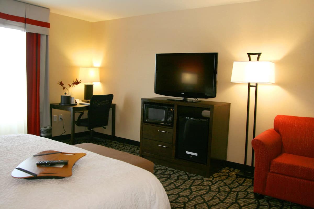 Een hotelkamer met een bed, een bureau met een computer, een televisie, een magnetron, een rode fauteuil en een lamp.