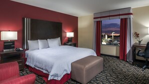 Quarto de hotel com uma cama grande, duas mesas de cabeceira com luminárias, uma poltrona vermelha, uma mesinha com um vaso e vista para a cidade pela janela.