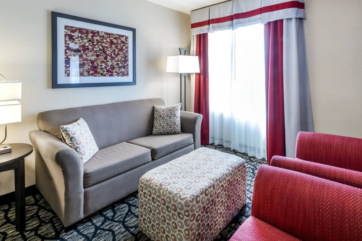 Hotelkamer met een bank, poef, fauteuil, een lamp, een lijst met kunstwerk en een raam met gordijnen.