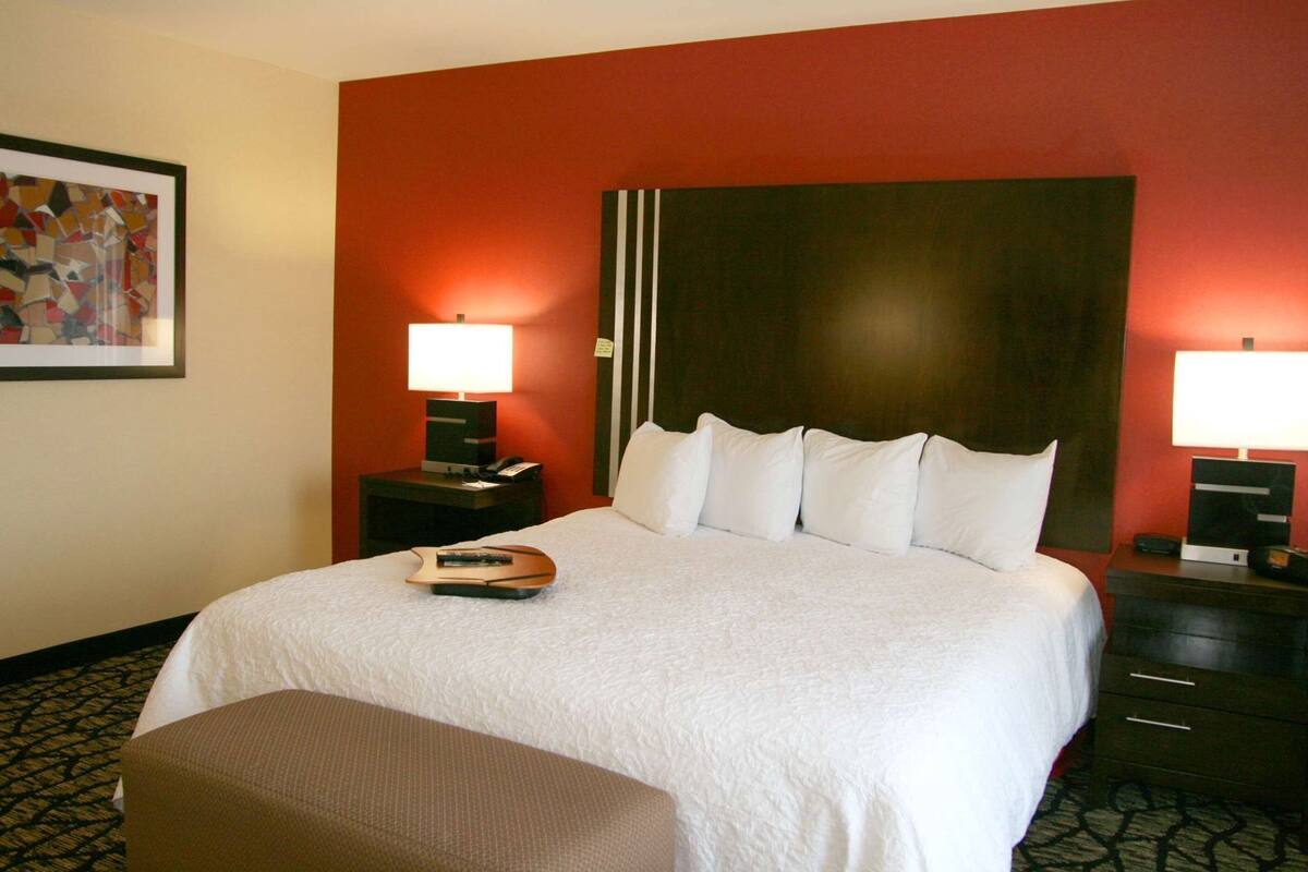 Une chambre d’hôtel avec un grand lit, deux tables de chevet avec des lampes, une télécommande sur le lit et un tableau encadré au mur.