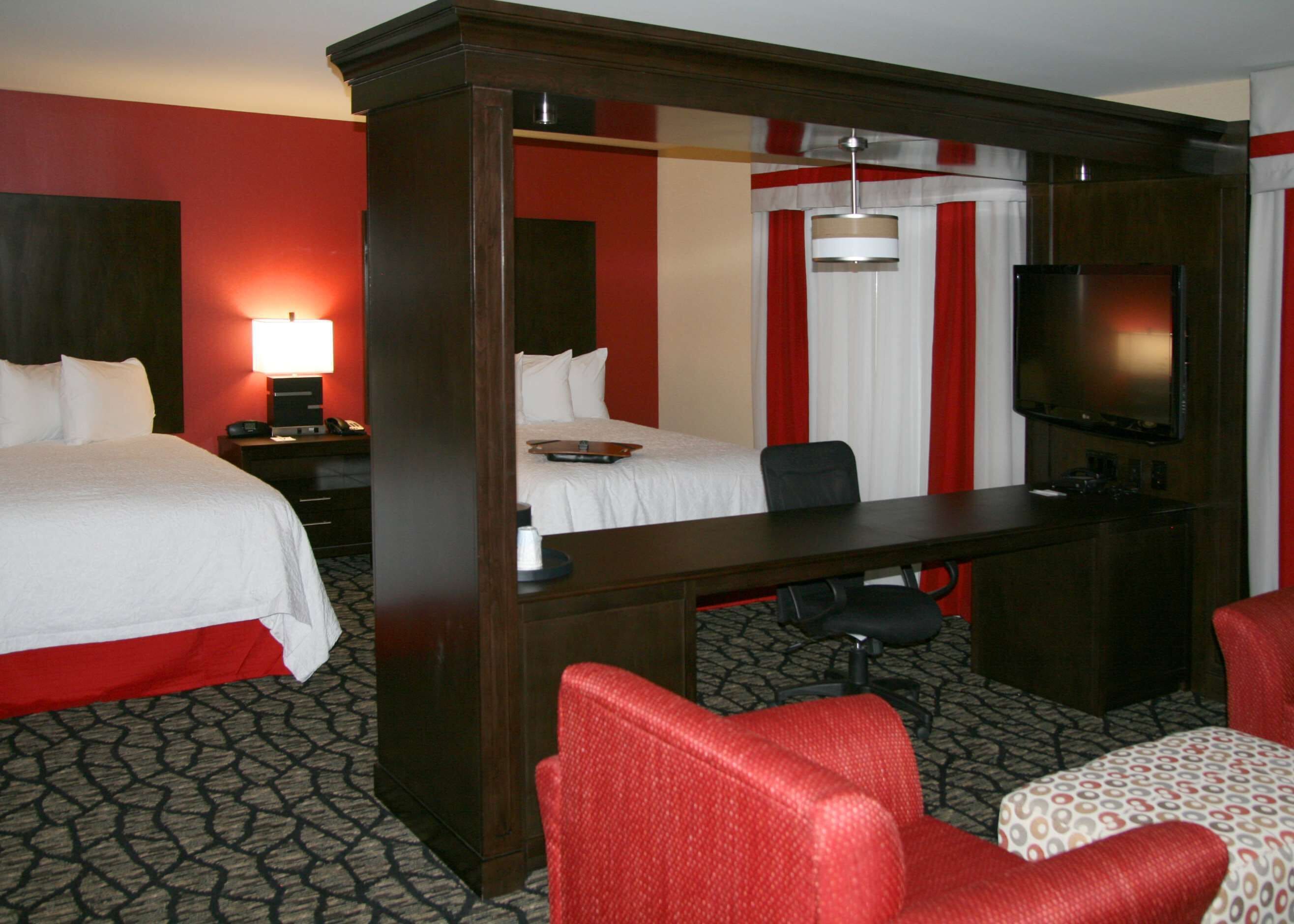 Ein Hotelzimmer mit einem großen Bett, einem Schreibtisch mit Stuhl, einem Fernseher und einem kleinen Tisch mit gemustertem Tischläufer.