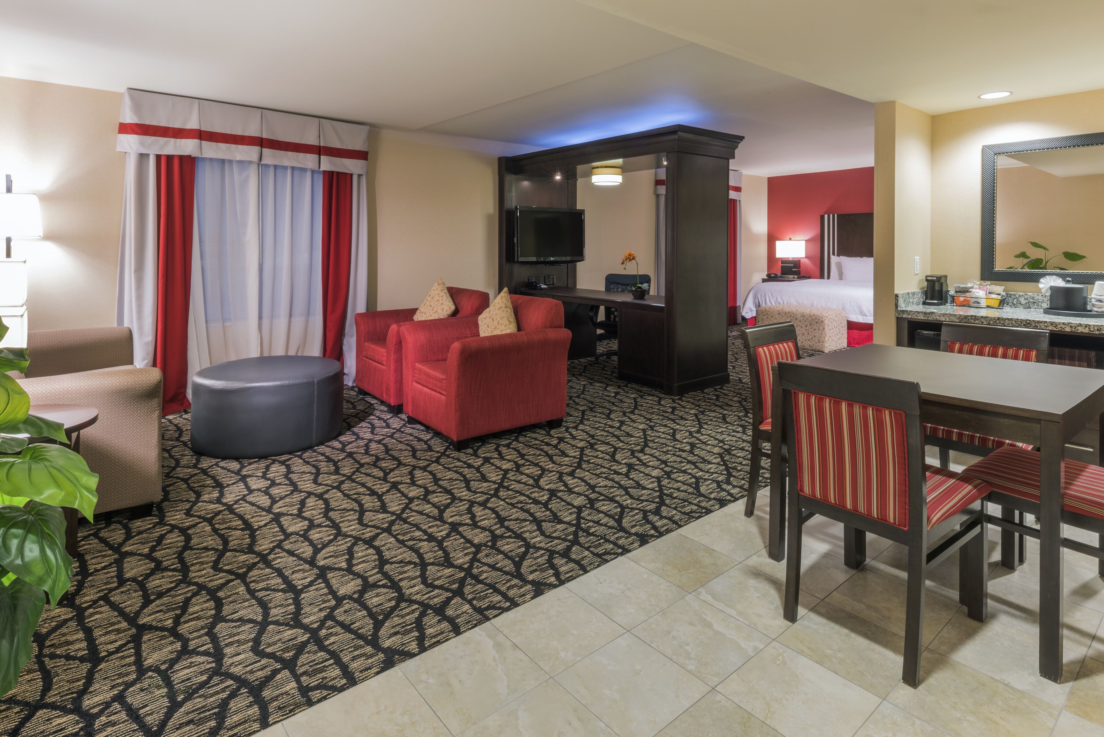 A hotel room with a living area featuring a red sofa, a round ottoman, a flat-screen TV, a dining table with chairs, and a kitchen area with a microwave and coffee maker.